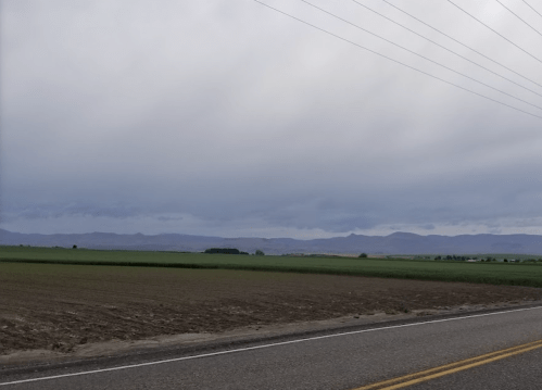 A wide view of a rural landscape with fields, distant mountains, and a cloudy sky above. Power lines run along the road.