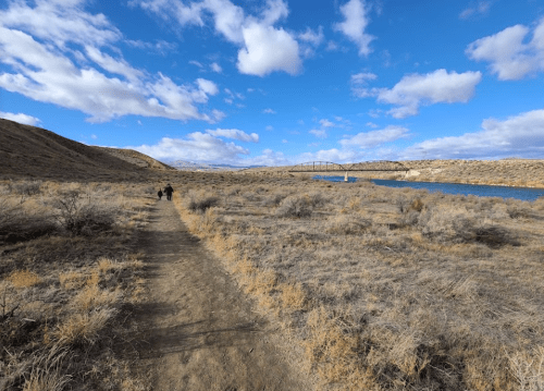 Two people walk along a dirt path beside a river, with a bridge in the distance and a blue sky filled with clouds.