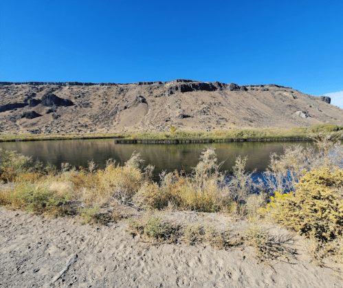 A serene river flows beside a rocky hillside, surrounded by sparse vegetation under a clear blue sky.