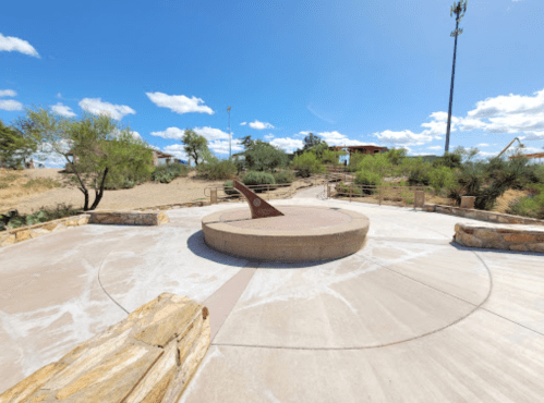 A sundial in a landscaped park with desert plants and a blue sky, surrounded by stone pathways and seating areas.