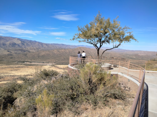 Two people stand on a lookout point, overlooking a vast mountainous landscape under a clear blue sky.