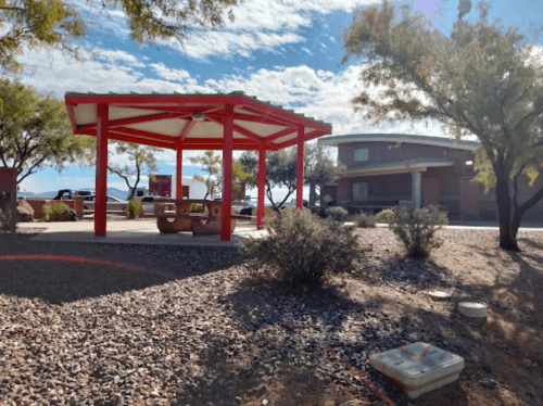 A red gazebo with picnic tables in a desert landscape, surrounded by shrubs and a building in the background.