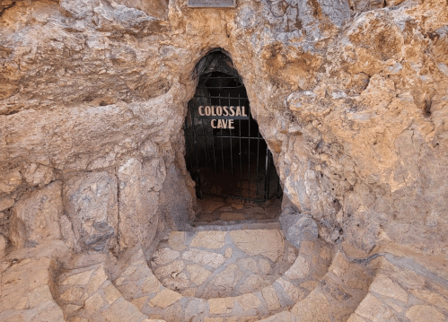 Entrance to Colossal Cave, featuring a stone pathway and a gated opening surrounded by rocky walls.