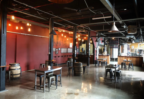 A spacious brewery interior with wooden barrels, metal tables, and warm lighting, featuring brewing equipment in the background.