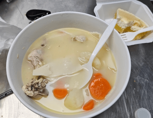 A bowl of soup with chicken, dumplings, carrots, and a white spoon, next to a dessert in a takeout container.