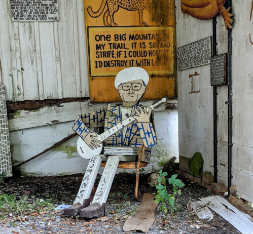 A wooden figure playing a banjo sits on a chair, surrounded by colorful signs and rustic decor.