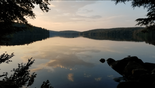 A serene lake at sunset, reflecting trees and clouds, surrounded by gentle hills and rocky shorelines.