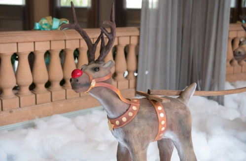 A decorative reindeer with antlers and a red nose, standing on a bed of faux snow indoors.