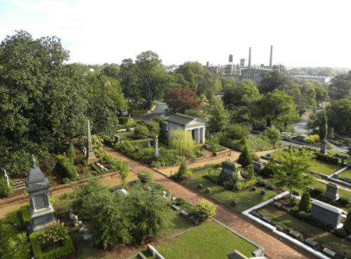 Aerial view of a cemetery with various gravestones and lush greenery, set against a backdrop of industrial buildings.