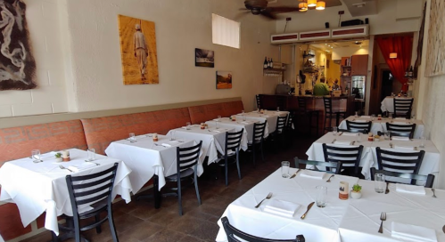 A cozy restaurant interior with white tablecloths, black chairs, and artwork on the walls, featuring a bar in the background.