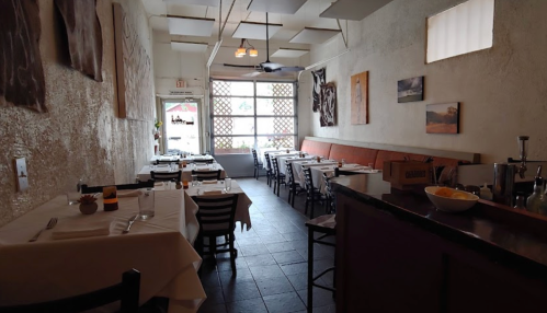 A cozy restaurant interior with white walls, tables set for dining, and large windows letting in natural light.