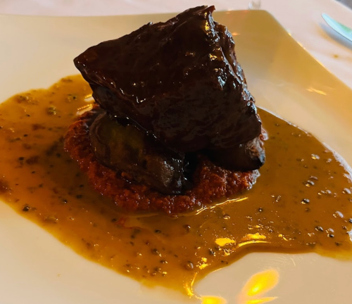 A plated dish featuring a large piece of meat in a rich sauce, served on a bed of grain with a drizzle of sauce.