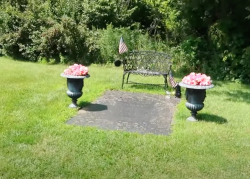 A peaceful memorial site featuring a bench, flower pots, and a small American flag, surrounded by greenery.