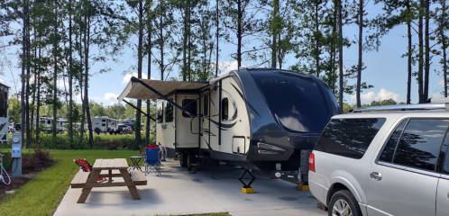 A parked RV with an awning in a campsite surrounded by trees, with a picnic table and a vehicle nearby.