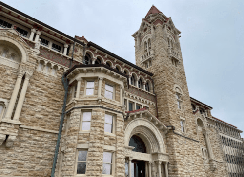 Historic stone building with intricate architectural details and a tall tower, set against a cloudy sky.