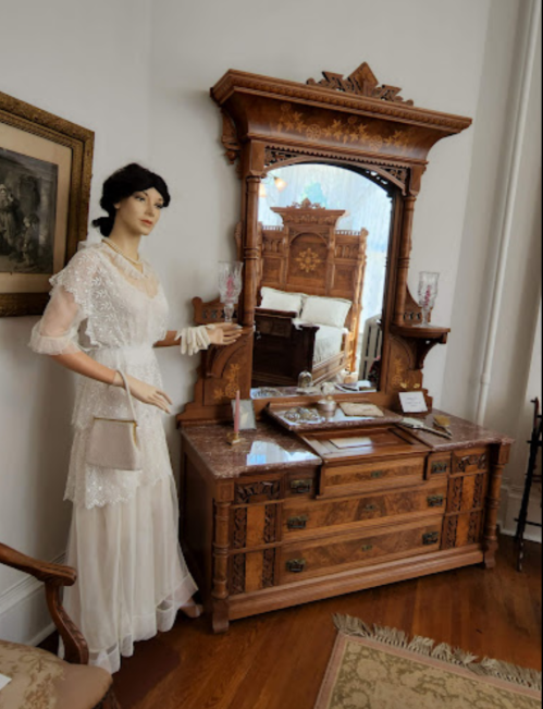 A vintage mannequin in a white dress stands beside an ornate wooden dresser and mirror in a museum setting.