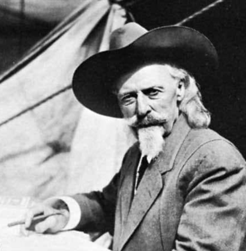A man with a mustache and long hair, wearing a large hat and suit, sits outdoors near a tent, holding a pen.