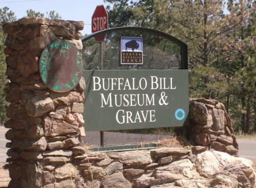 Sign for the Buffalo Bill Museum and Grave, featuring a stone structure and a stop sign in the background.