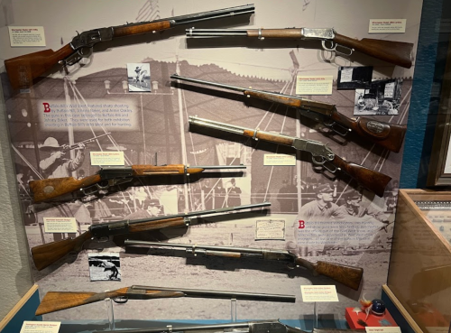Display of historical rifles and shotguns mounted on a wall, with informative labels and a vintage photo backdrop.