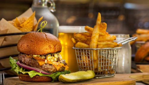 A juicy burger with cheese, lettuce, and onion, served with fries and a pickle, alongside a glass of beer.