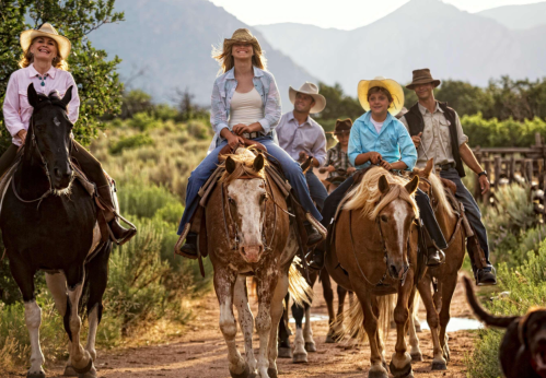 A group of six people on horseback, wearing cowboy hats, riding along a scenic trail with mountains in the background.