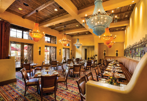 Elegant restaurant interior with colorful chandeliers, wooden beams, and a patterned carpet, featuring neatly set tables.