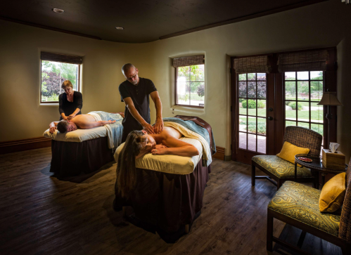 Two people receiving massages in a serene spa room, with a third therapist attending to one of them. Natural light filters in.