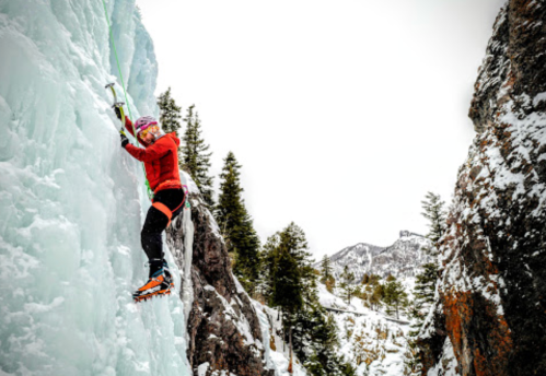 A climber ascends an icy wall in a snowy landscape, surrounded by trees and rocky mountains.