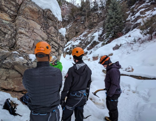 Four climbers in helmets prepare for ice climbing in a snowy, rocky area surrounded by trees.