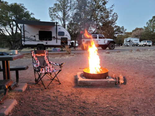 A campfire burns brightly in a fire pit, with camping chairs nearby and RVs parked in the background under a twilight sky.