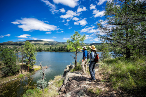 Two hikers stand on a rocky outcrop, overlooking a serene lake surrounded by trees and mountains under a blue sky.
