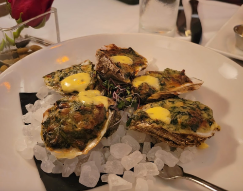 A plate of baked oysters topped with a creamy sauce, served on crushed ice with a garnish of microgreens.
