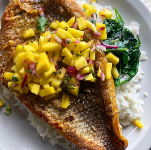 A plate of fried fish topped with mango salsa, served on a bed of rice with sautéed greens.