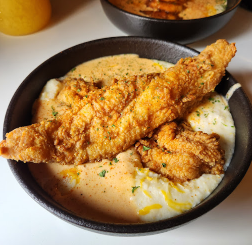 A bowl of creamy grits topped with fried fish fillets and garnished with herbs and spices.