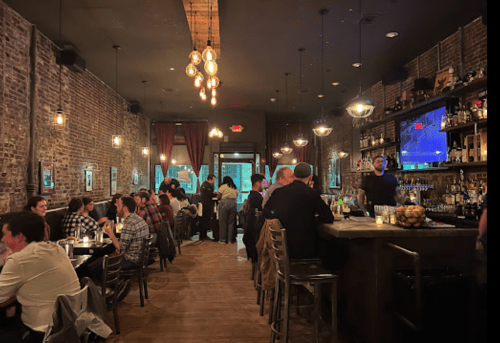 A lively restaurant interior with exposed brick walls, dim lighting, and patrons enjoying drinks and conversation at tables.