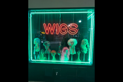 A brightly lit shop window displaying various wigs and a neon "WIGS" sign.