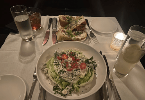 A dinner table set with a salad topped with tomatoes, two pieces of bread, and drinks in dim lighting.