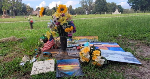A memorial site with flowers, paintings, and a sign expressing gratitude, set in a grassy area.