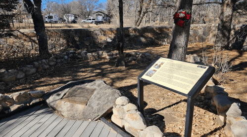 A stone plaque and informational sign in a wooded area, with a wreath hanging on a nearby tree.