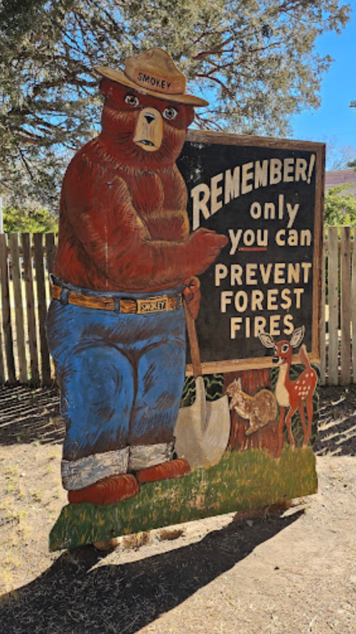 A wooden sign featuring Smokey Bear, reminding viewers to prevent forest fires, with a shovel and deer nearby.