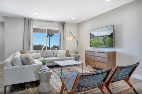 Modern living room with a gray sofa, two woven chairs, a coffee table, and a TV displaying a sports event.