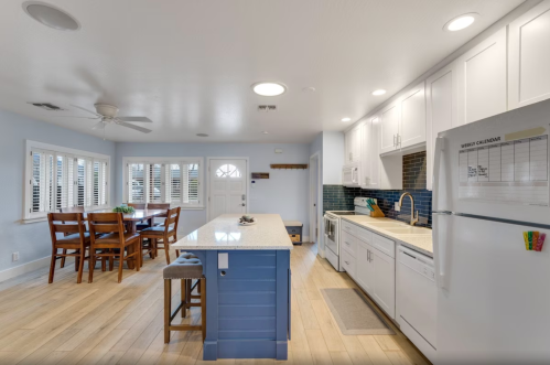 Bright kitchen with white cabinets, blue accents, a dining table, and large windows, featuring modern appliances and wood flooring.