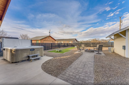 A backyard with a hot tub, fire pit, seating area, and green lawn under a blue sky with scattered clouds.