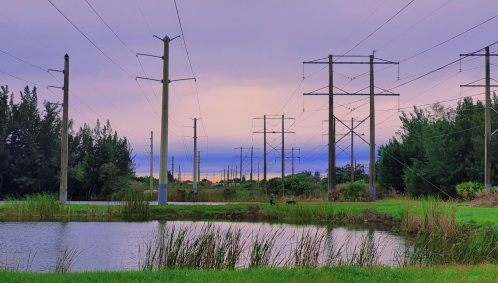 Power lines stretch across a landscape with a pond and greenery under a colorful sky at dusk.