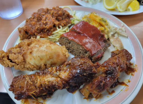 A plate of comfort food featuring fried chicken, meatloaf, barbecue ribs, spaghetti, and sides.