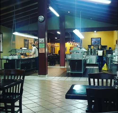 A spacious restaurant interior with metal serving stations, tables, and staff preparing food in a warm, inviting atmosphere.