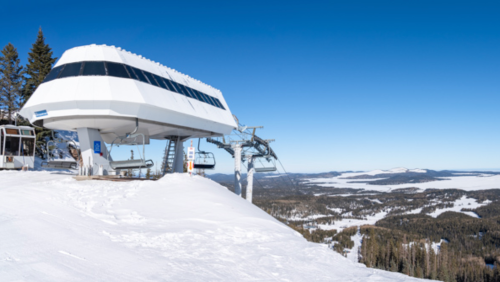 Ski lift station on a snowy mountain with clear blue skies and a panoramic view of the landscape below.