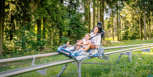 A woman and a child enjoy a ride on a mountain coaster through a lush, green forest.