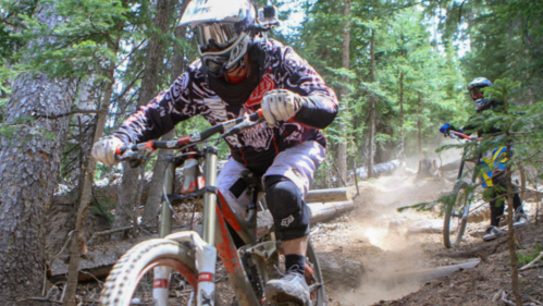 Two mountain bikers navigate a dusty trail through a forest, surrounded by trees and dirt.
