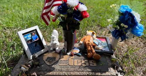 A grave marker adorned with flowers, a flag, photos, and small figurines, set in a grassy area.